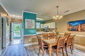 Enjoy meals in this dining area with a large table and six chairs, surrounded by a cityscape painting and a kitchen with white cabinets.