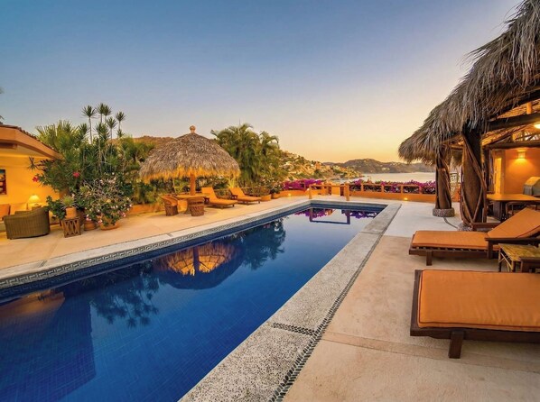 A swimming pool with lounge chairs and a thatched-roof structure, surrounded by palm trees and a clear sky. - Rarely Available- Casa Quixote - Beachfront Estate (Zihuatanjeo)