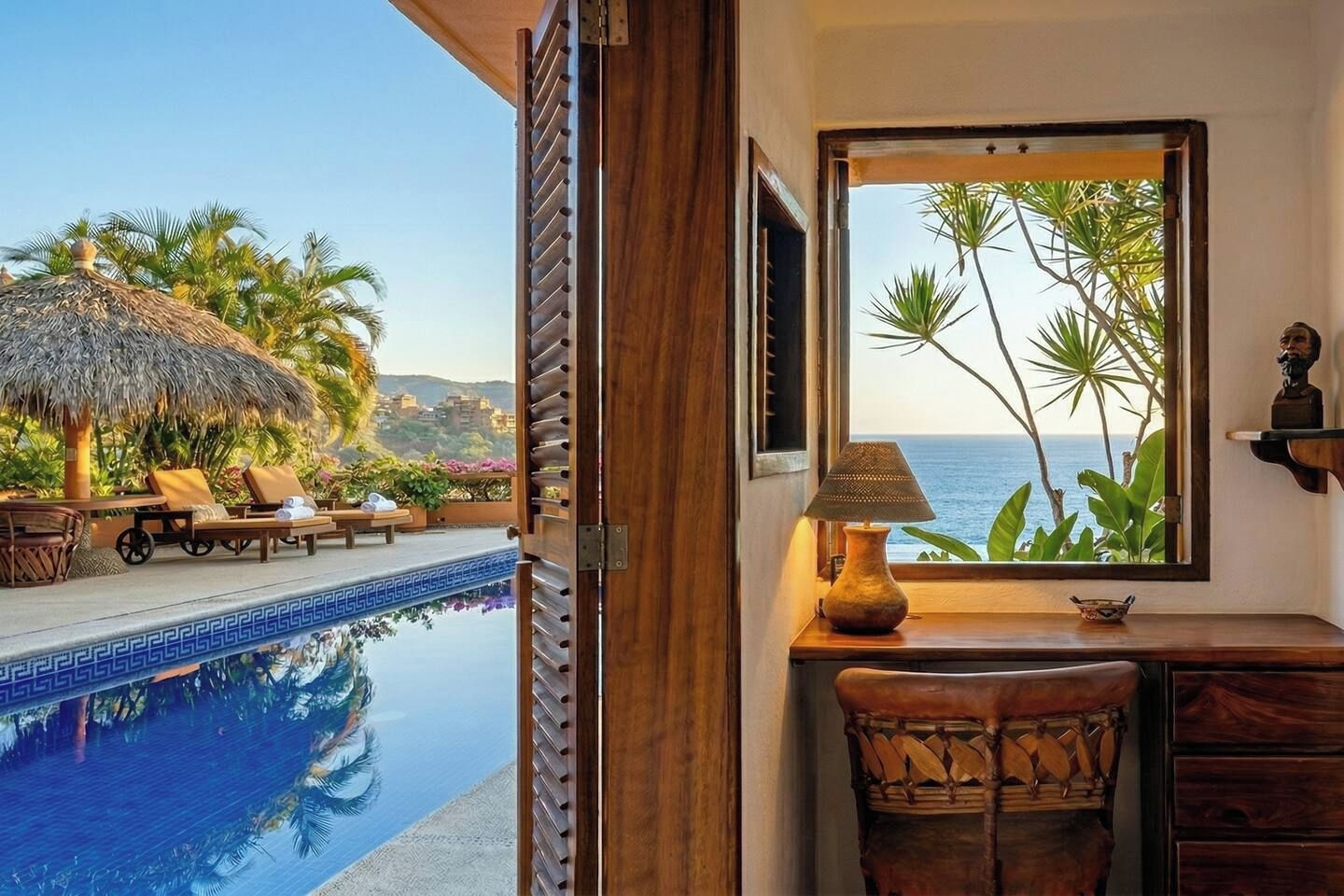Habitación con vista a una piscina y palmeras, un escritorio de madera con una lámpara y una silla.