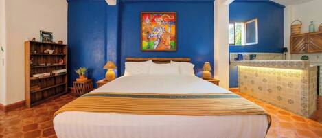 A bedroom with a large bed, a wooden headboard, a blue accent wall, a painting, a wooden cabinet, and a tiled floor.