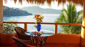 A balcony with a table, chairs, a vase of flowers, and a view of the sea and hills.