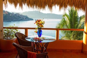 A balcony with a table, chairs, a vase of flowers, and a view of the sea and hills.