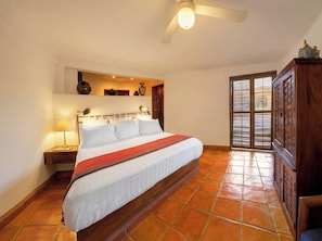Un dormitorio con una cama grande, una mesita de noche, una lámpara, un ventilador de techo y vistas a través de una puerta.