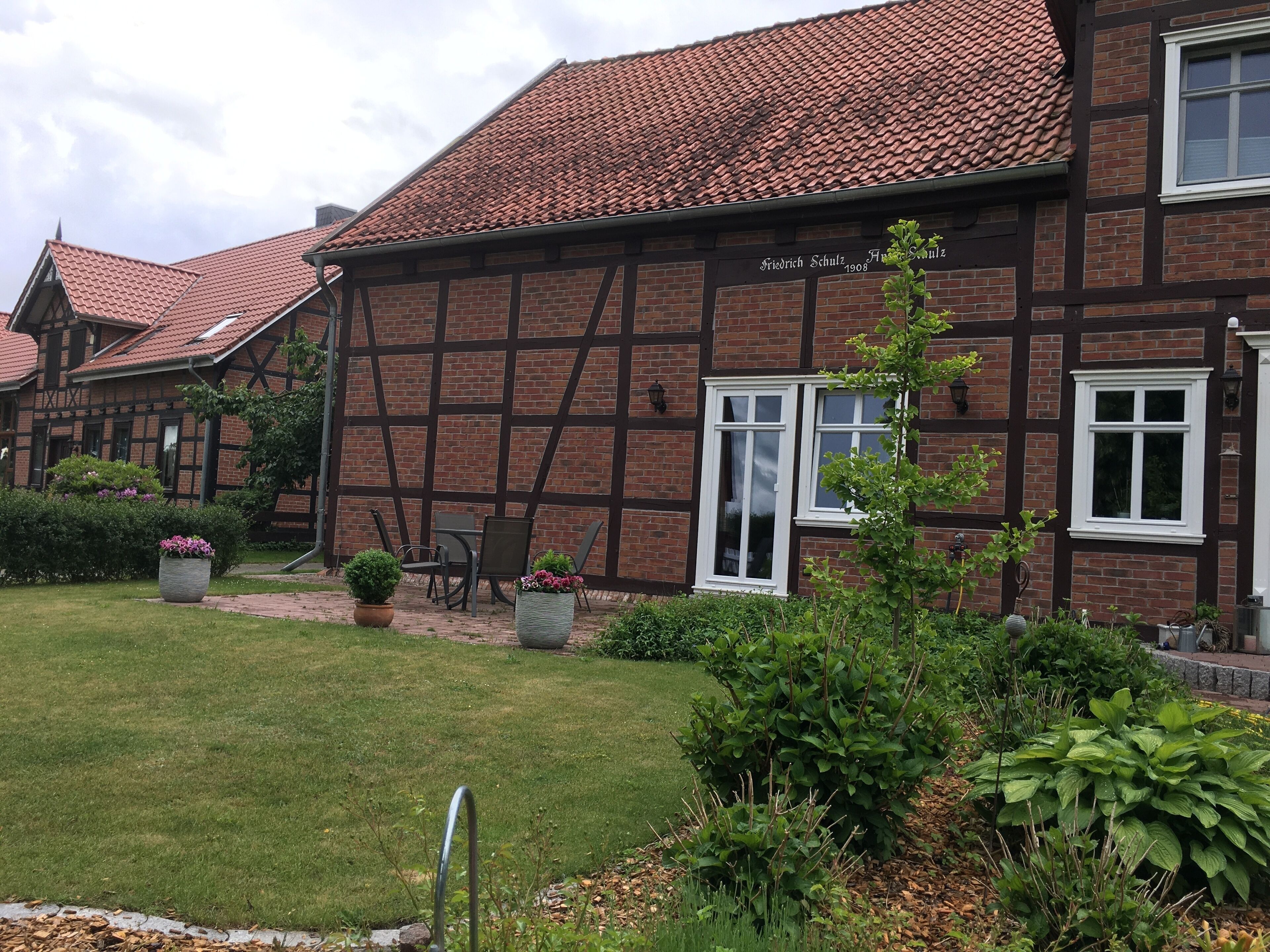 Un bâtiment traditionnel à colombages, avec un toit recouvert de tuiles rouges, une plaque portant son nom et un jardin bien entretenu agrémenté de plantes en pot et d’une pelouse.