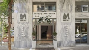 Exterior de um hotel com fachada de mármore, um letreiro com a inscrição HOTEL APARTMENTS MENI e uma escadaria que leva à entrada.