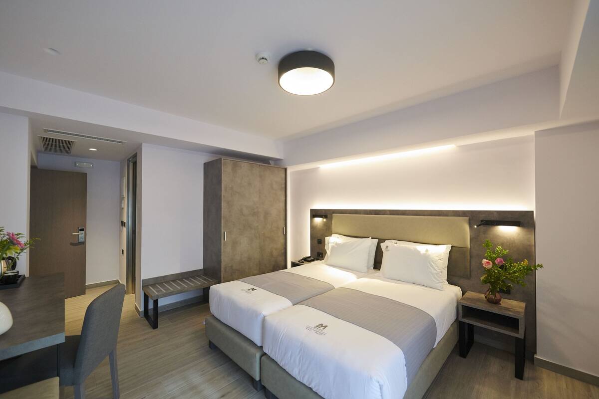 Ein Hotelzimmer mit Doppelbett, Schreibtisch mit Stuhl, einer Bank, Nachttisch mit Lampe und einer kleinen Topfpflanze.