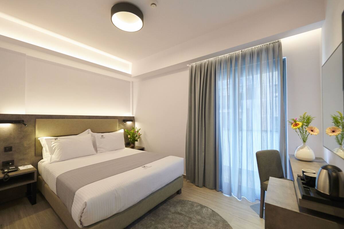Ein modernes Hotelzimmer mit einem großen Bett, Nachttischen, einem Schreibtisch mit Stuhl, einer Vase mit Blumen und einem Fenster mit Vorhängen.
