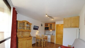 A compact living space with a kitchenette, wooden furniture, a TV, and a window with curtains. - Cozy studio with balcony, ski-in/ski-out (Les Menuires)