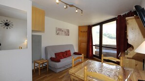 A cozy room with a wooden floor, a grey sofa, a wooden dining table with chairs, a wooden cabinet, and a window with curtains. - Cozy studio with balcony, ski-in/ski-out (Les Menuires)