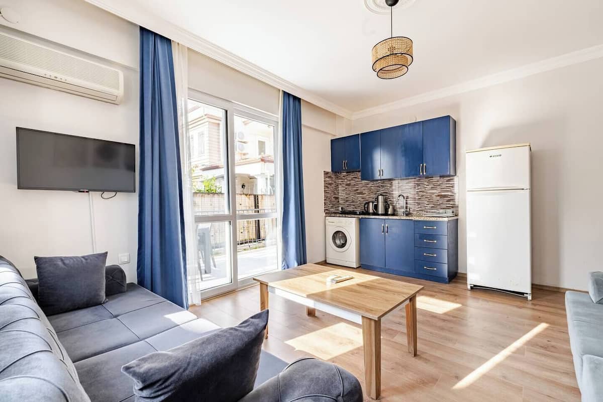 Una sala moderna con un sofá, una mesa de centro y un televisor. Se observa una cocina con armarios azules y una lavadora.
