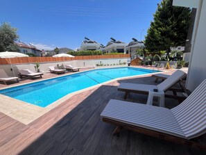 A swimming pool with lounge chairs, wooden deck, and a view of houses in the background.