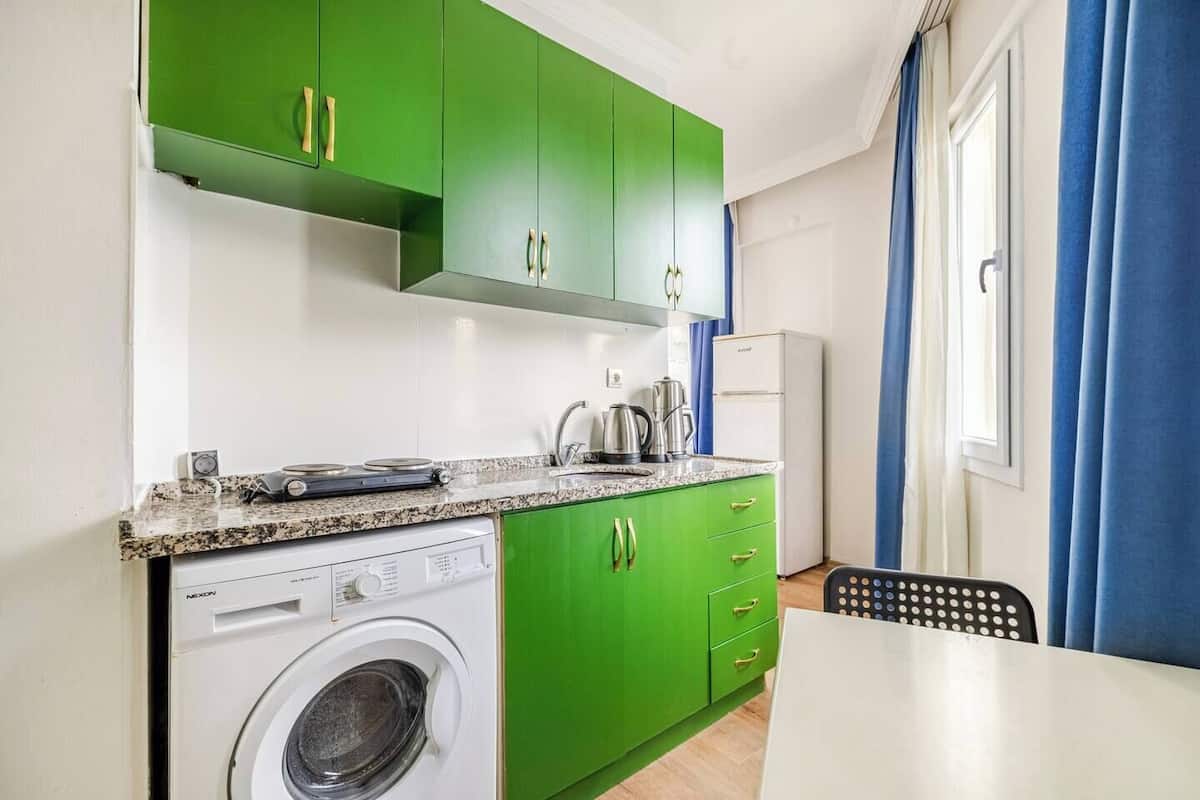 Una cocina compacta con armarios verdes, lavadora, cafetera y un comedor con mesa y sillas.