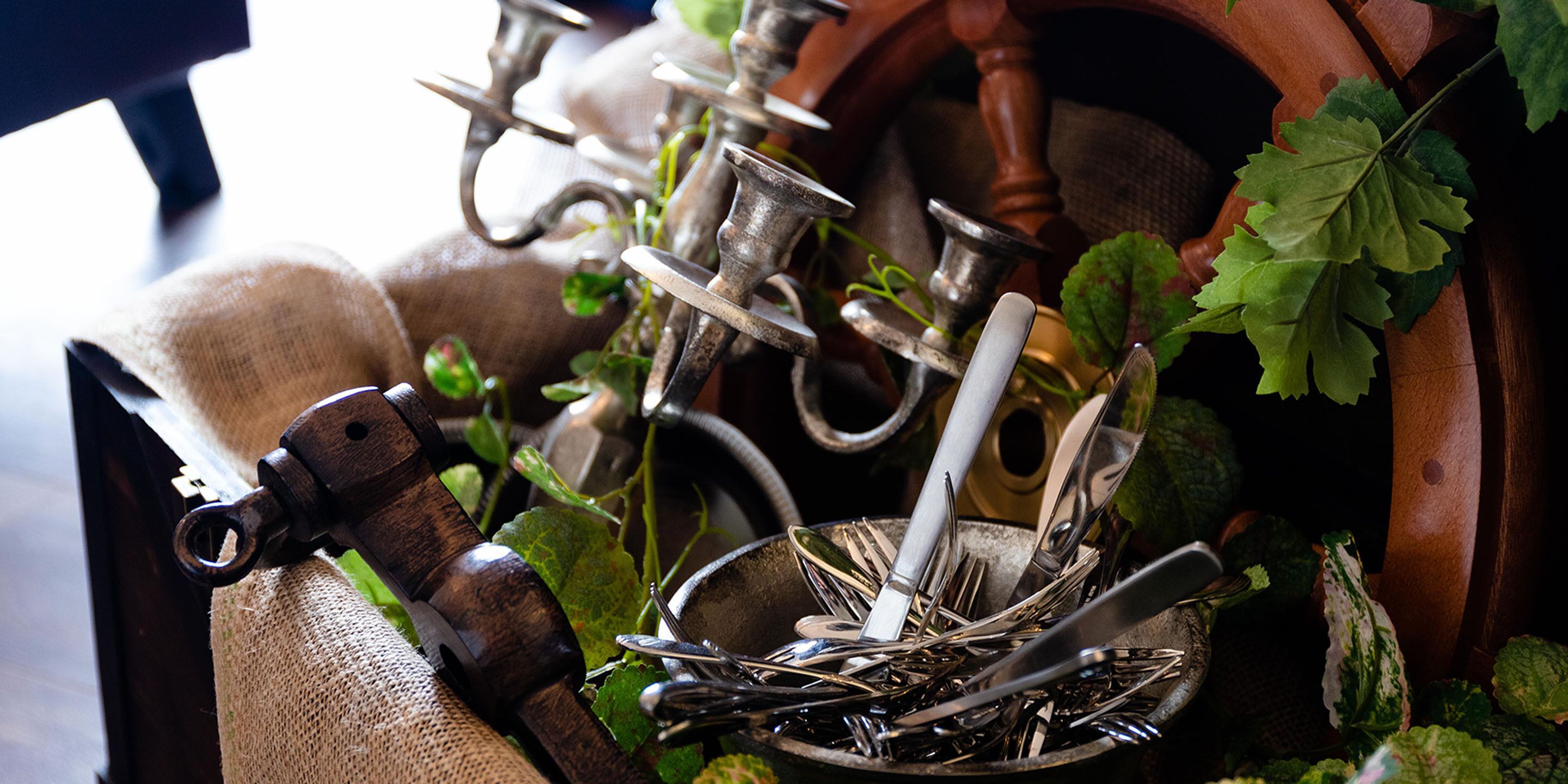 A collection of antique kitchen utensils, including forks, knives, and spoons, arranged in a rustic setting with a burlap sack and green leaves.