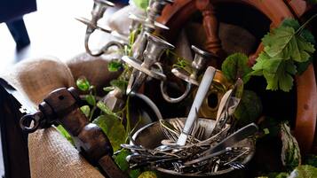 A collection of antique kitchen utensils, including forks, knives, and spoons, arranged in a rustic setting with a burlap sack and green leaves.