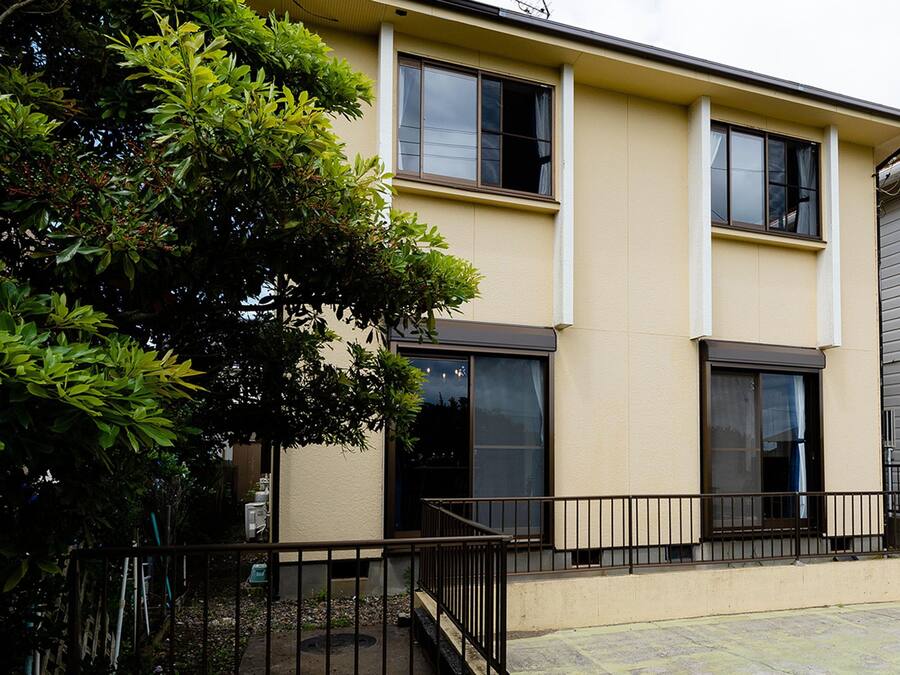 A two-story residential building with a beige exterior, large windows, and a black metal railing in front.