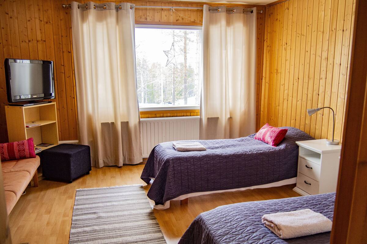 Una habitación con dos camas, un televisor en una estantería, una ventana con cortinas y una pared de madera.