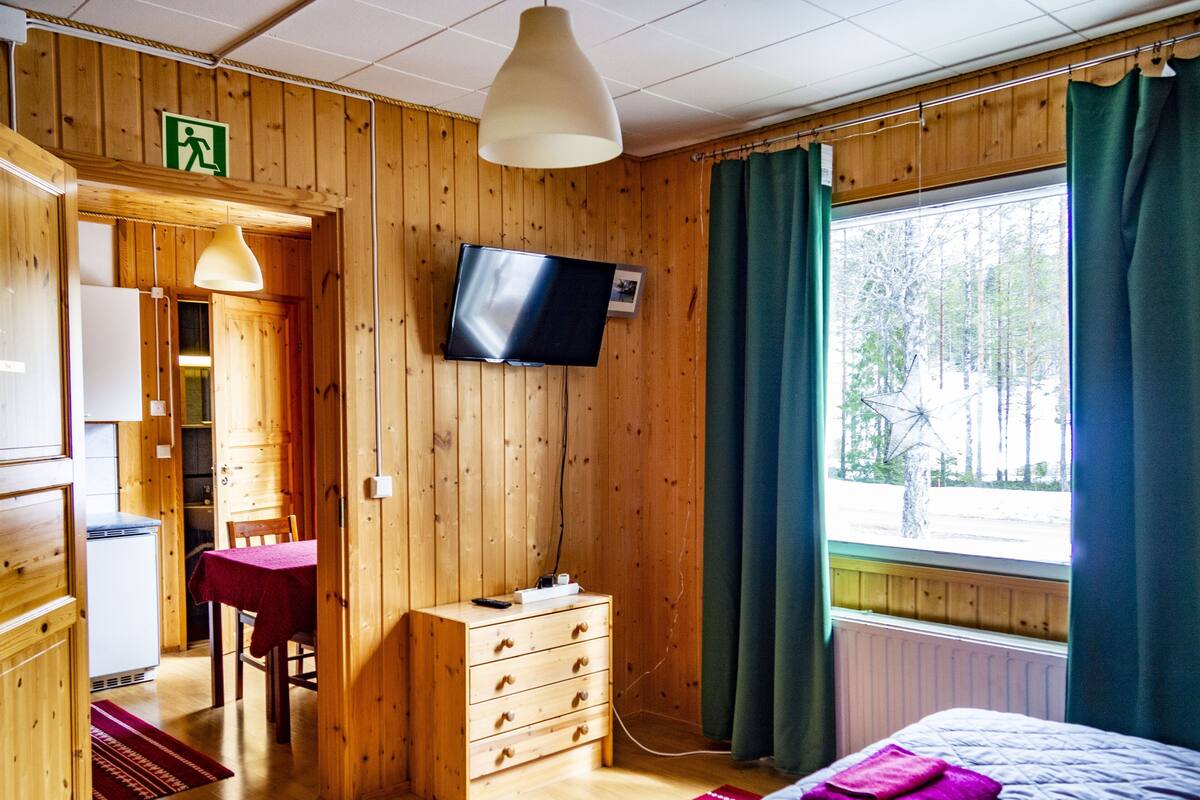 Una habitación de madera con una cama, un aparador, un escritorio con silla, un televisor y una ventana con cortinas.