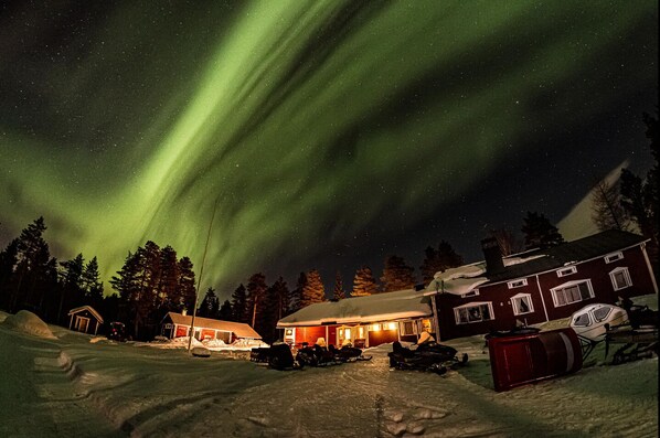 A snow-covered landscape with a building and snowmobiles, under a night sky with the Northern Lights.