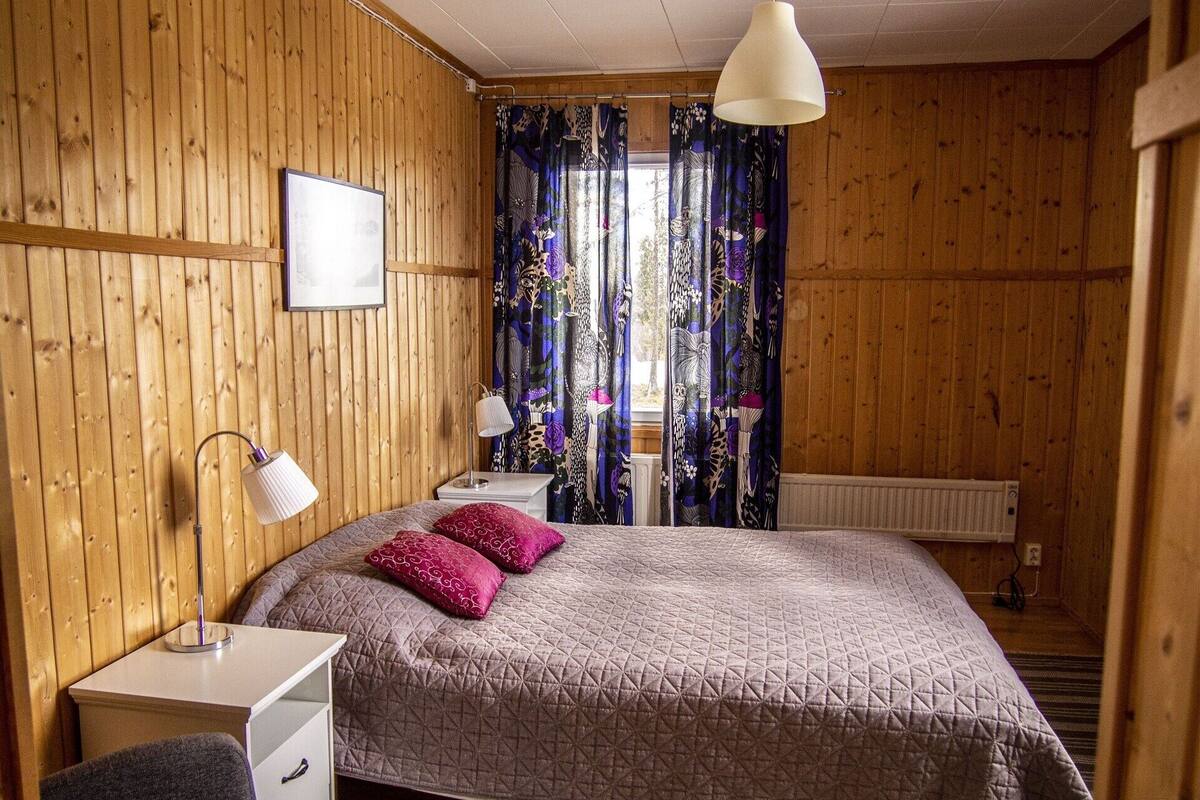 Un dormitorio con paredes de madera, una cama con colcha acolchada, una mesita de noche con lámpara y una ventana con cortinas florales.