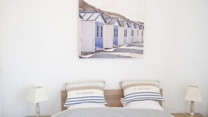A bedroom with a bed, two bedside lamps, a framed picture of beach huts, and two pillows with text.