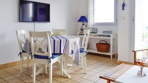 A dining area with a table set for four, a television mounted on the wall, a window with curtains, and a small desk with a lamp and a basket.