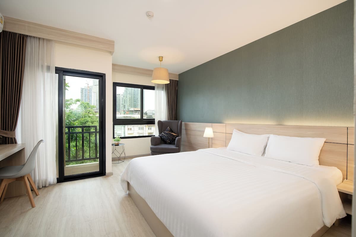 A hotel room with a large bed, a desk, a chair, a lamp, and a view of the city through a window.