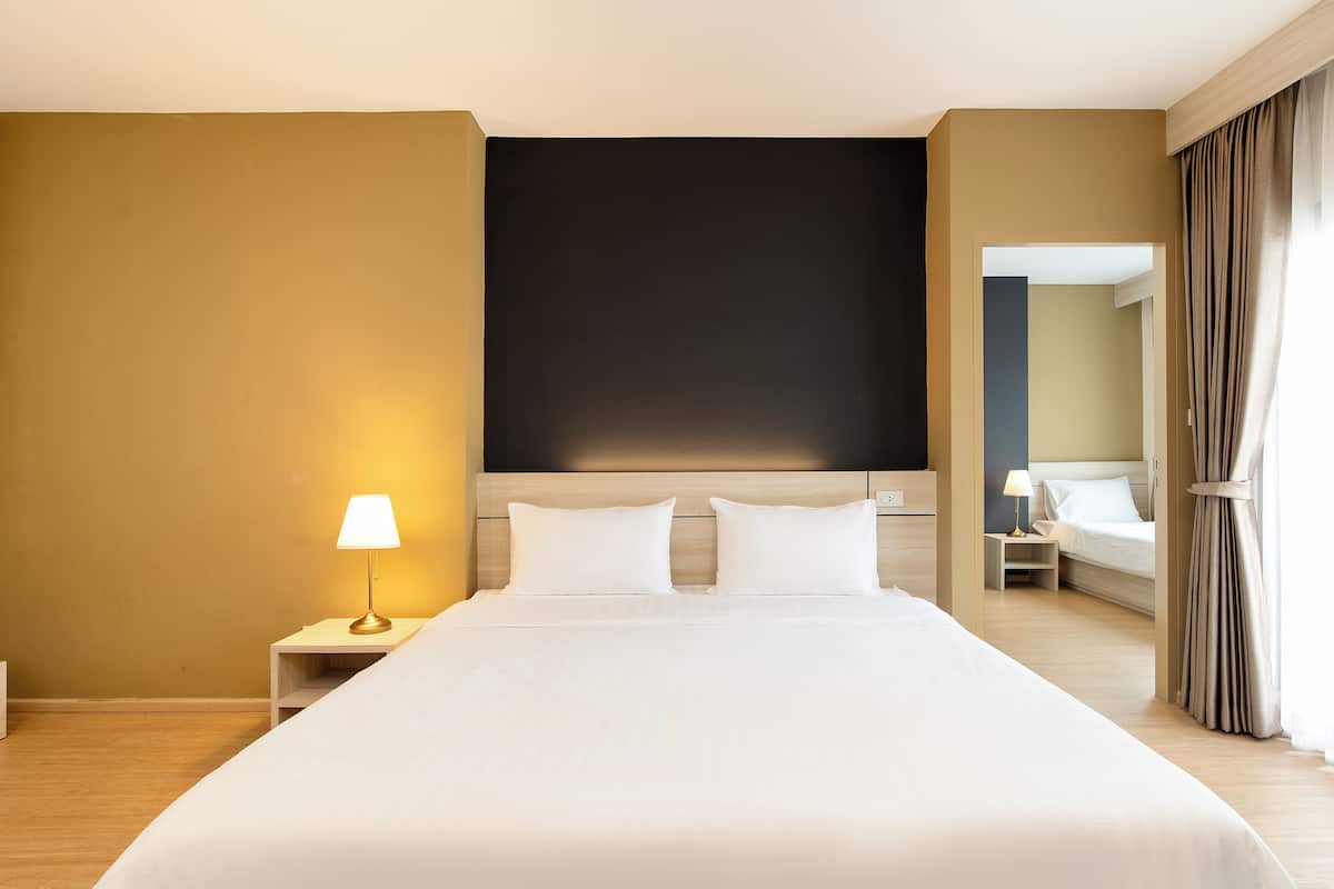 A hotel room with a large bed, a nightstand with a lamp, a dark headboard, and a view of another room through a doorway.