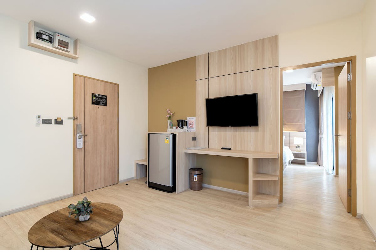 A compact hotel room with a kitchenette, a flat-screen TV mounted on the wall, a small table with a plant, and a door with a digital lock.