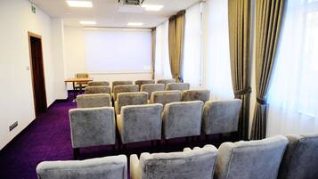 Un salón de conferencias con filas de sillas grises, una pantalla de proyector y cortinas en las ventanas.
