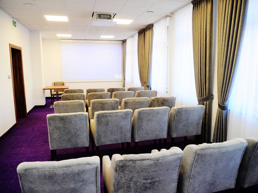 Un salón de conferencias con filas de sillas grises, una pantalla de proyector y cortinas en las ventanas.