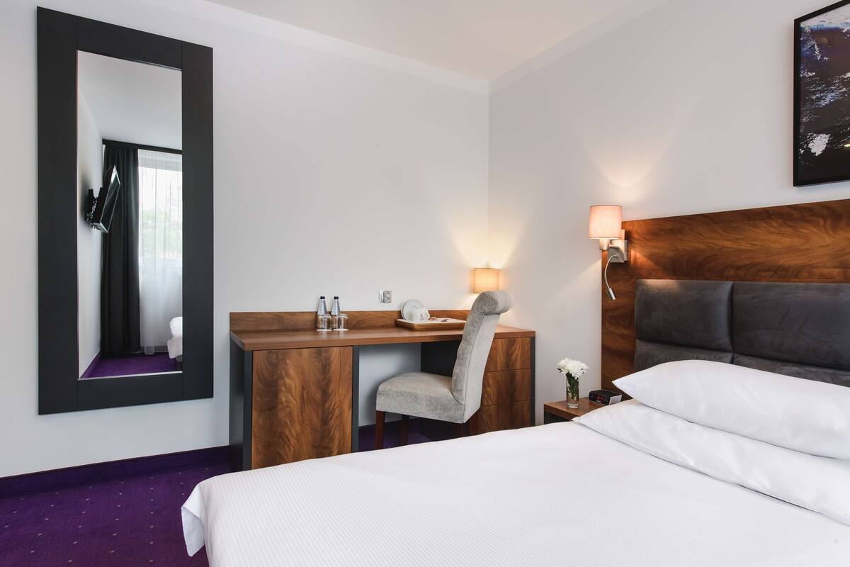 A hotel room with a bed, a desk with a chair, a mirror, a nightstand with a lamp, and a wall-mounted picture.