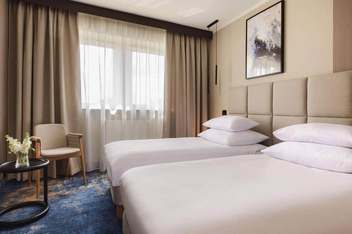 A hotel room with two beds, a chair, a small table with a vase of flowers, and a painting on the wall.