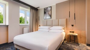 Una habitación de hotel moderna con una cama grande, una mesita de noche, una obra de arte en la pared y vistas a árboles por la ventana.