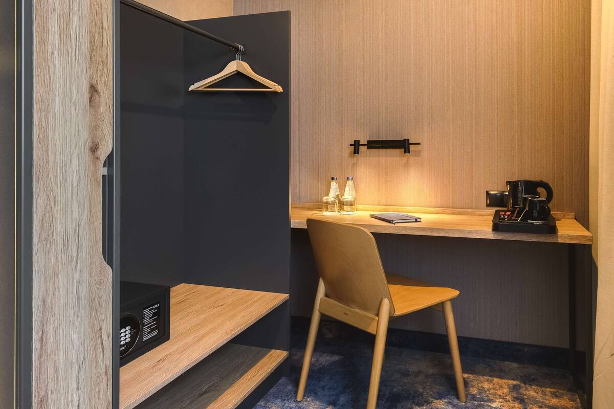 A modern hotel room with a wooden desk, a chair, a hanging coat rack, and a small shelf with bottles.