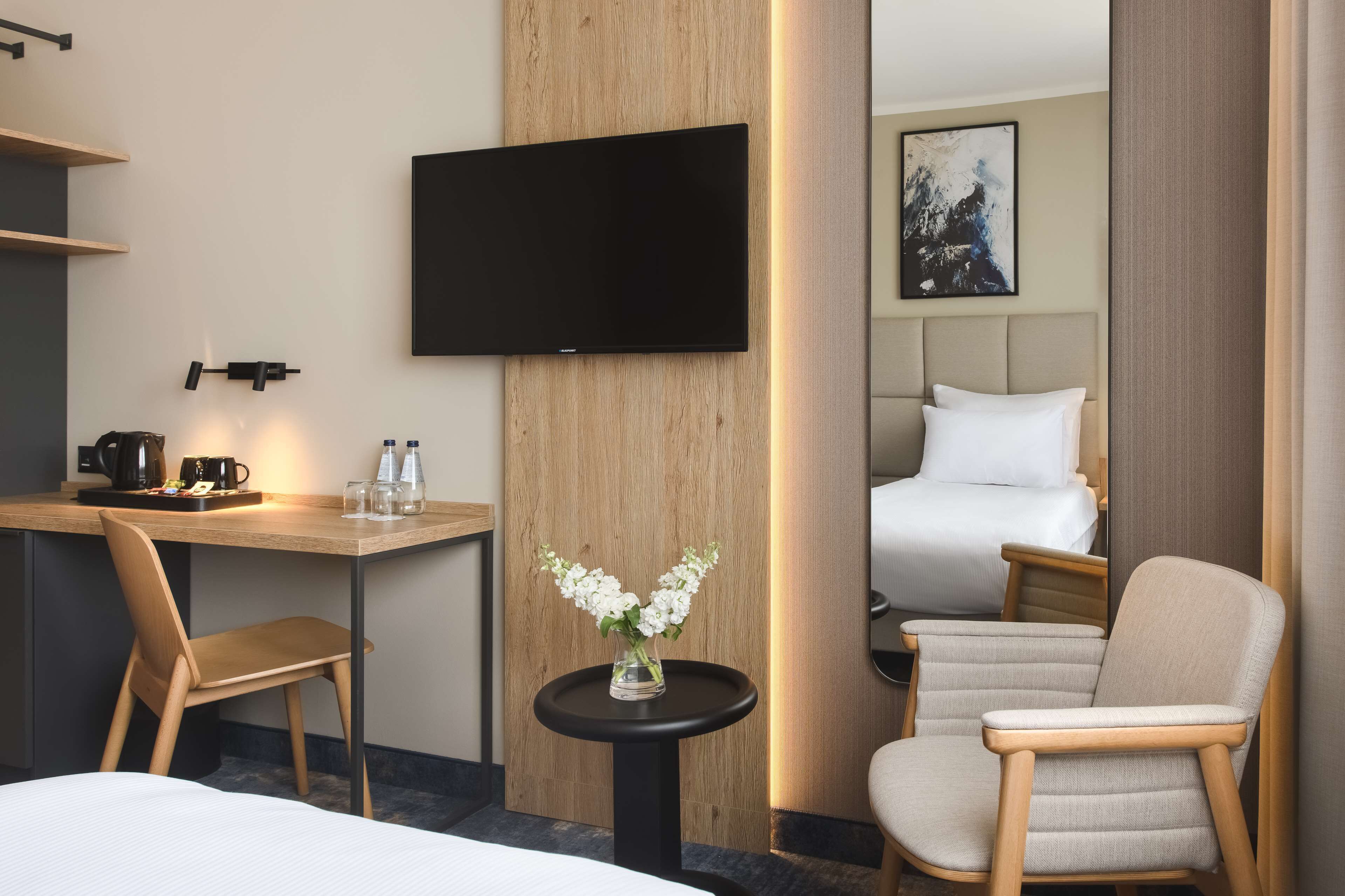 A hotel room with a bed, a desk with a coffee maker, a TV mounted on the wall, a small table with a vase of flowers, and a chair.