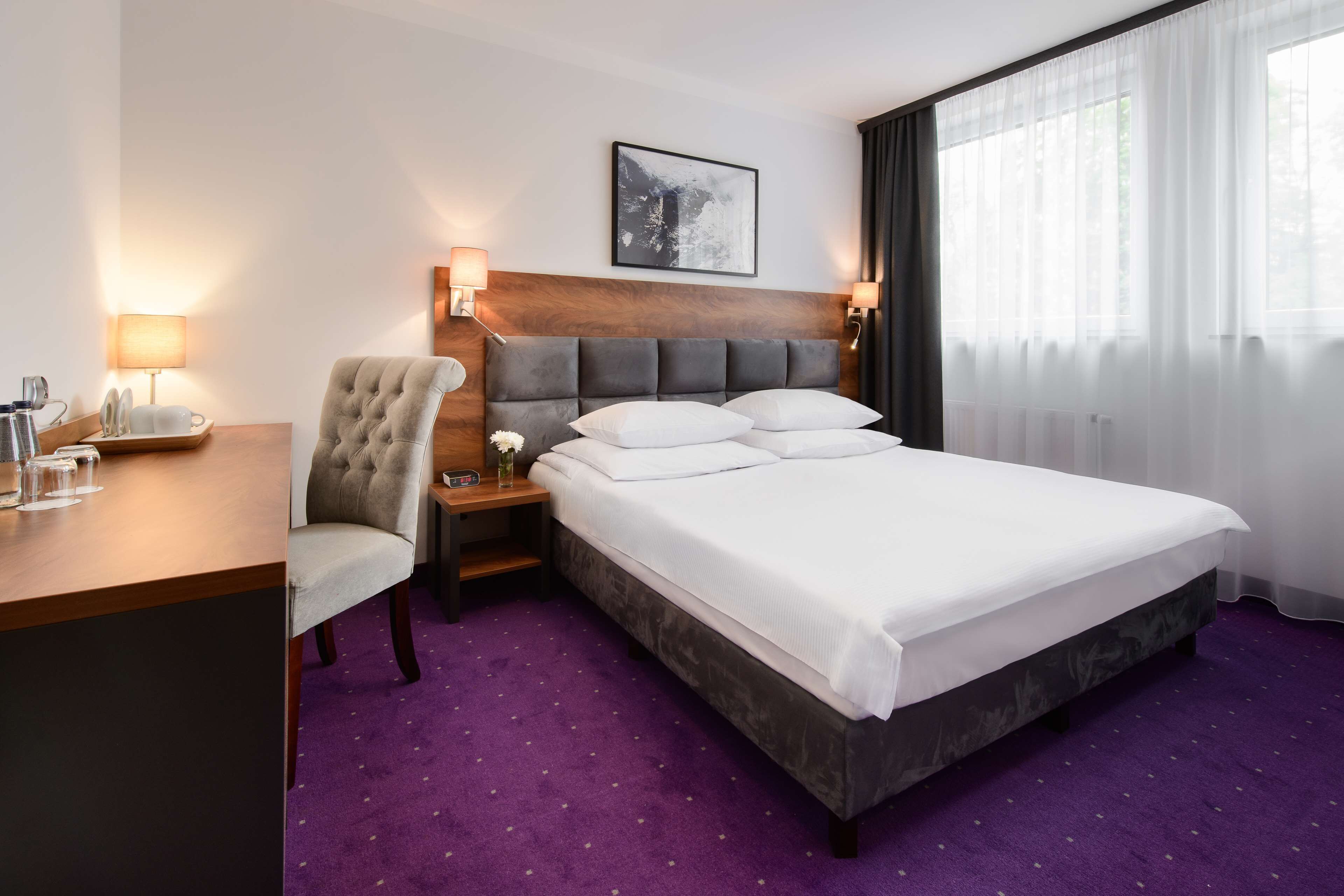 Habitació d'hotel amb un llit gran, una cadira, un escriptori amb una làmpada i una cambrera de nit amb una làmpada.