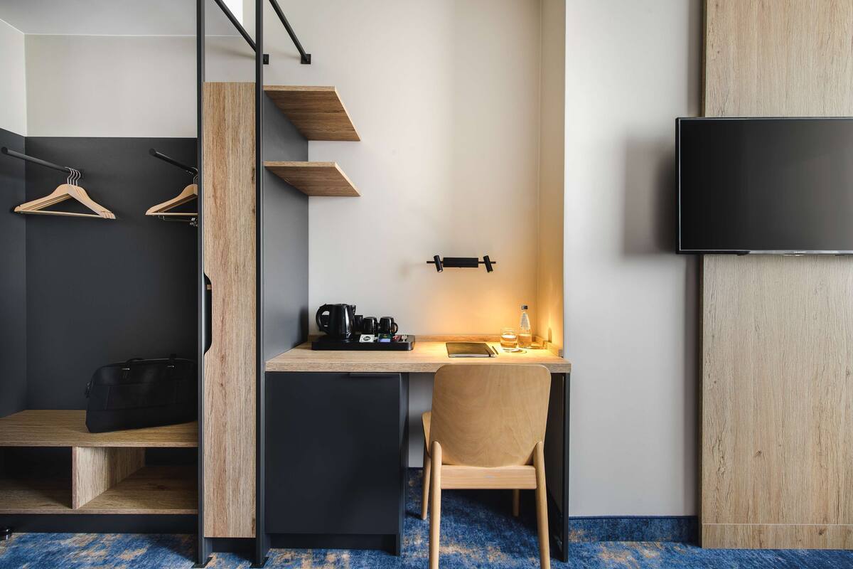 A compact hotel room with a wooden desk, a chair, a flat-screen TV, and a small seating area with a bench and a coat rack.