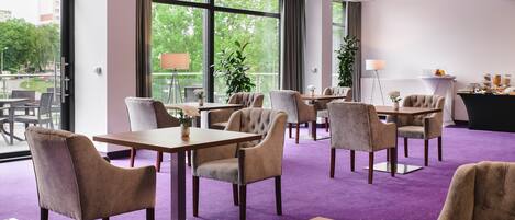 Un comedor de hotel con ventanas amplias, alfombra morada y varias mesas y sillas.