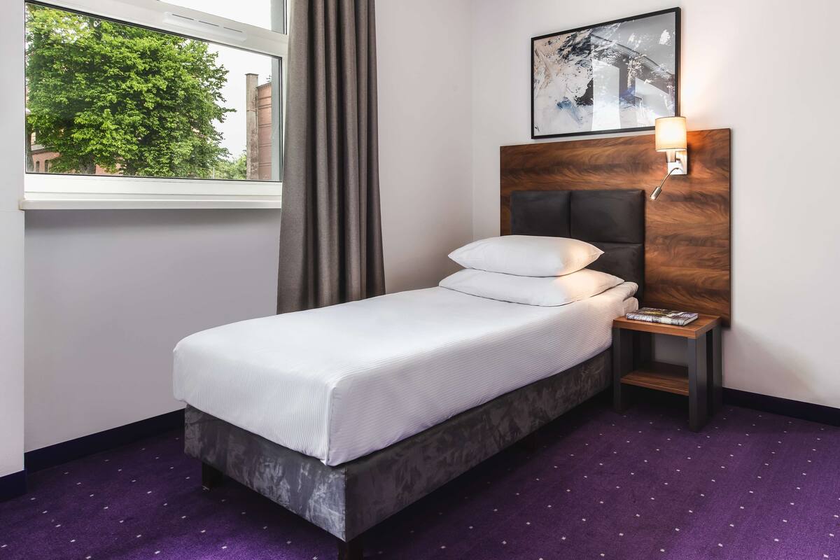 A single bed with a wooden headboard, a nightstand with a lamp, and a window with curtains.