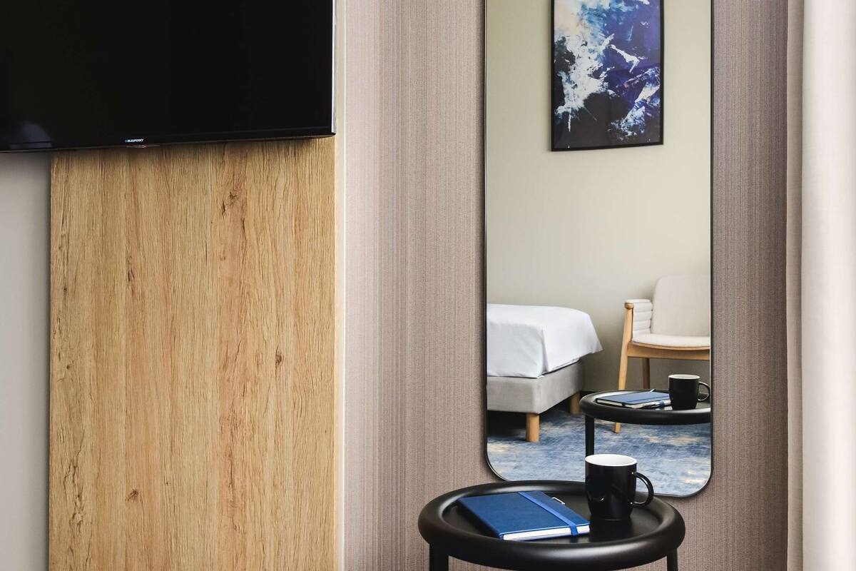 A hotel room with a wooden panel wall, a flat-screen TV mounted on it, a round black table with a blue notebook and a black mug, and a large mirror reflecting the room.