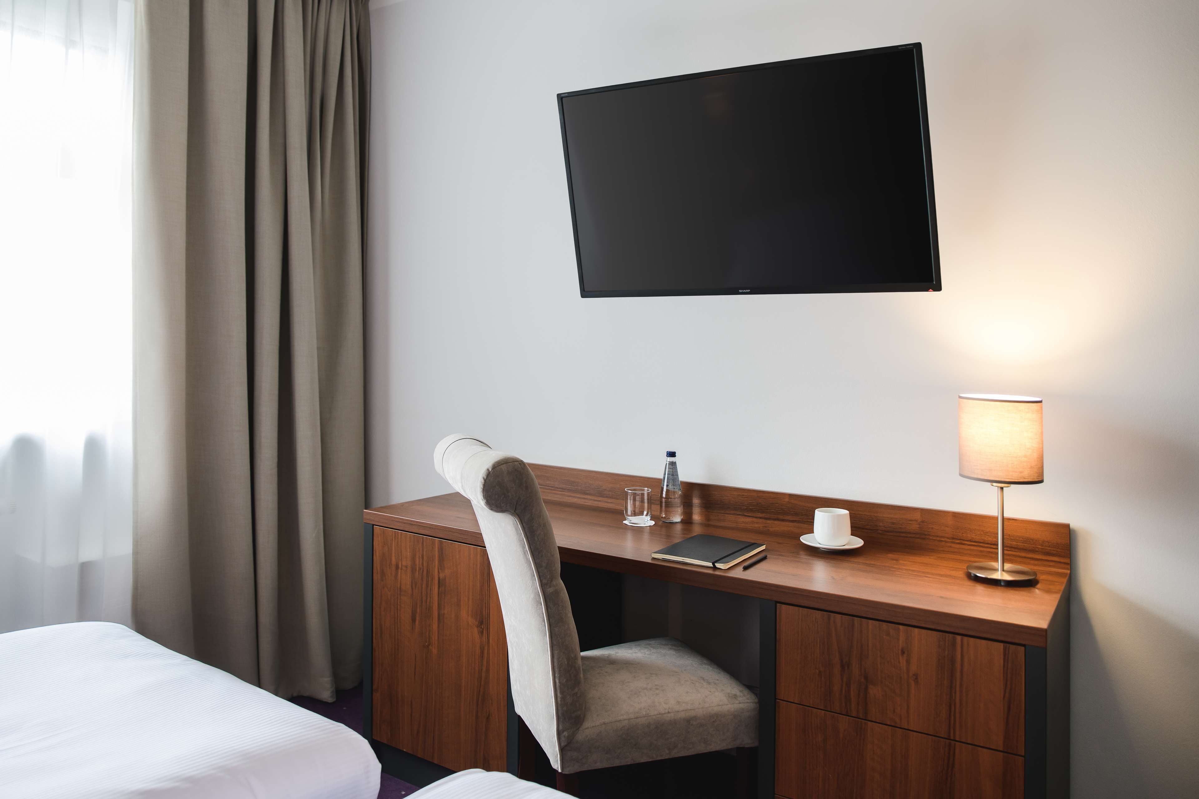 Une chambre d’hôtel comprenant un lit, un bureau avec une chaise, un téléviseur fixé au mur, une lampe et une table de chevet avec une tasse et une bouteille.