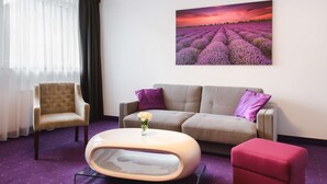 Una sala de estar moderna con un sofá color beige, un otomano rojo, una mesa de centro redonda y una silla. Un cuadro grande de campos de lavanda adorna la pared.