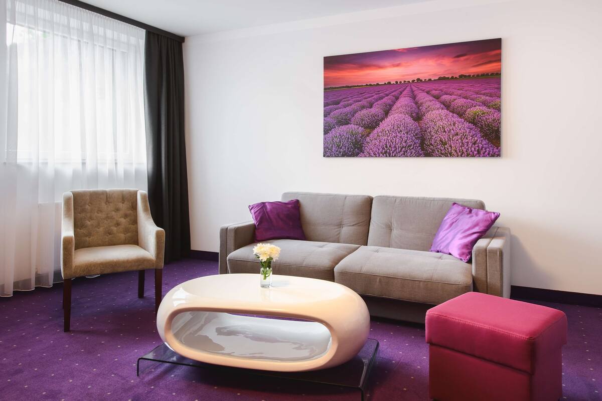 A modern living room with a beige sofa, a red ottoman, a round coffee table, and a chair. A large painting of lavender fields is on the wall.