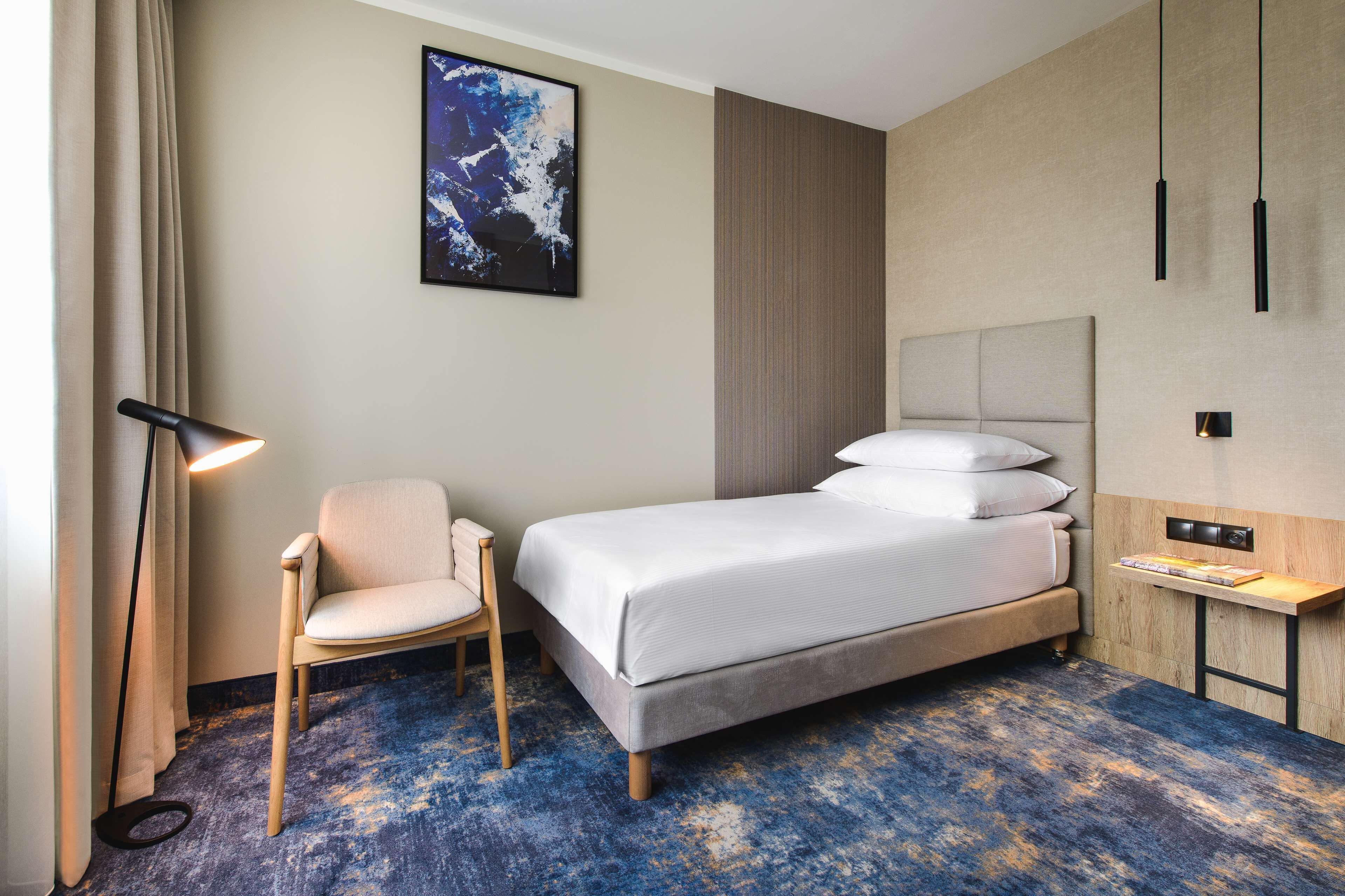 Een hotelkamer met een bed, een stoel, een bureau, een lamp en een schilderij aan de muur.