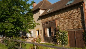 Un bâtiment rustique en pierre, doté d’une porte en bois et de volets aux fenêtres, entouré de verdure et délimité par une clôture en bois.