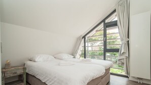 Un dormitorio con un ventanal grande, una cama con ropa de cama blanca, una mesita de noche de madera y un armario blanco.