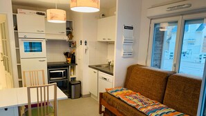 Une cuisine compacte avec un coin repas, un canapé agrémenté de coussins colorés et une fenêtre donnant sur des immeubles.