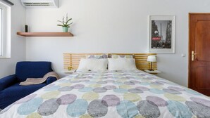Een slaapkamer met een bed, een blauwe fauteuil, een houten hoofdeinde, een wandairconditioner, een plank met een plant, een lijst met een afbeelding en een deur.