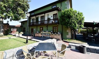 Un edificio de dos pisos con fachada verde, un balcón, zona de estar exterior con sillas de mimbre y una mesa de metal, y un pequeño jardín.