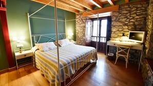 A four-poster bed with a striped bedspread, a stone wall, a wooden desk with a computer, and a window with a view. - Posada Término (Entrambasaguas)